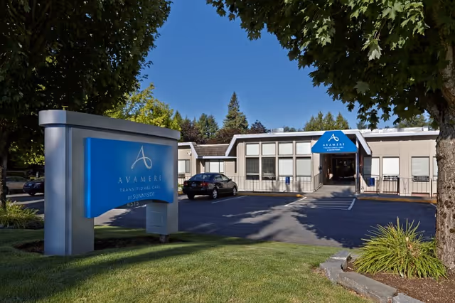 Exterior view of Avamere Transitional Care at Sunnyside building with a blue sign in the foreground displaying the facility name. The building is single-story with large windows and a blue awning over the entrance. There are trees and a parking lot with a car in front of the building under a clear blue sky.