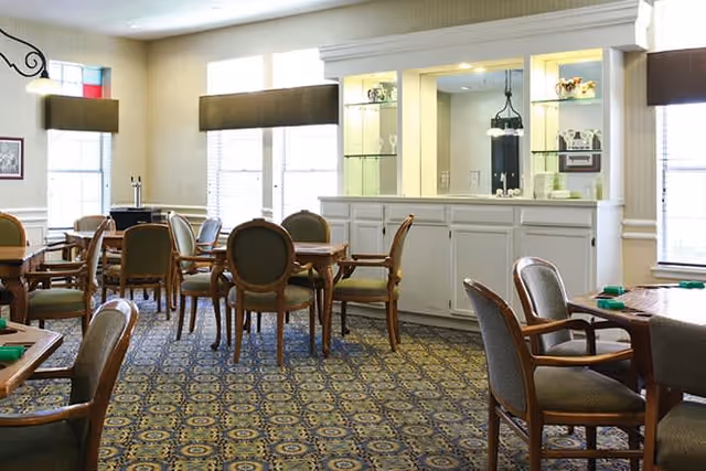 A well-lit dining room with multiple wooden tables and cushioned chairs arranged neatly on a patterned carpet. The room features large windows with brown valances, white cabinetry with glass shelves displaying decorative items, and soft overhead lighting.