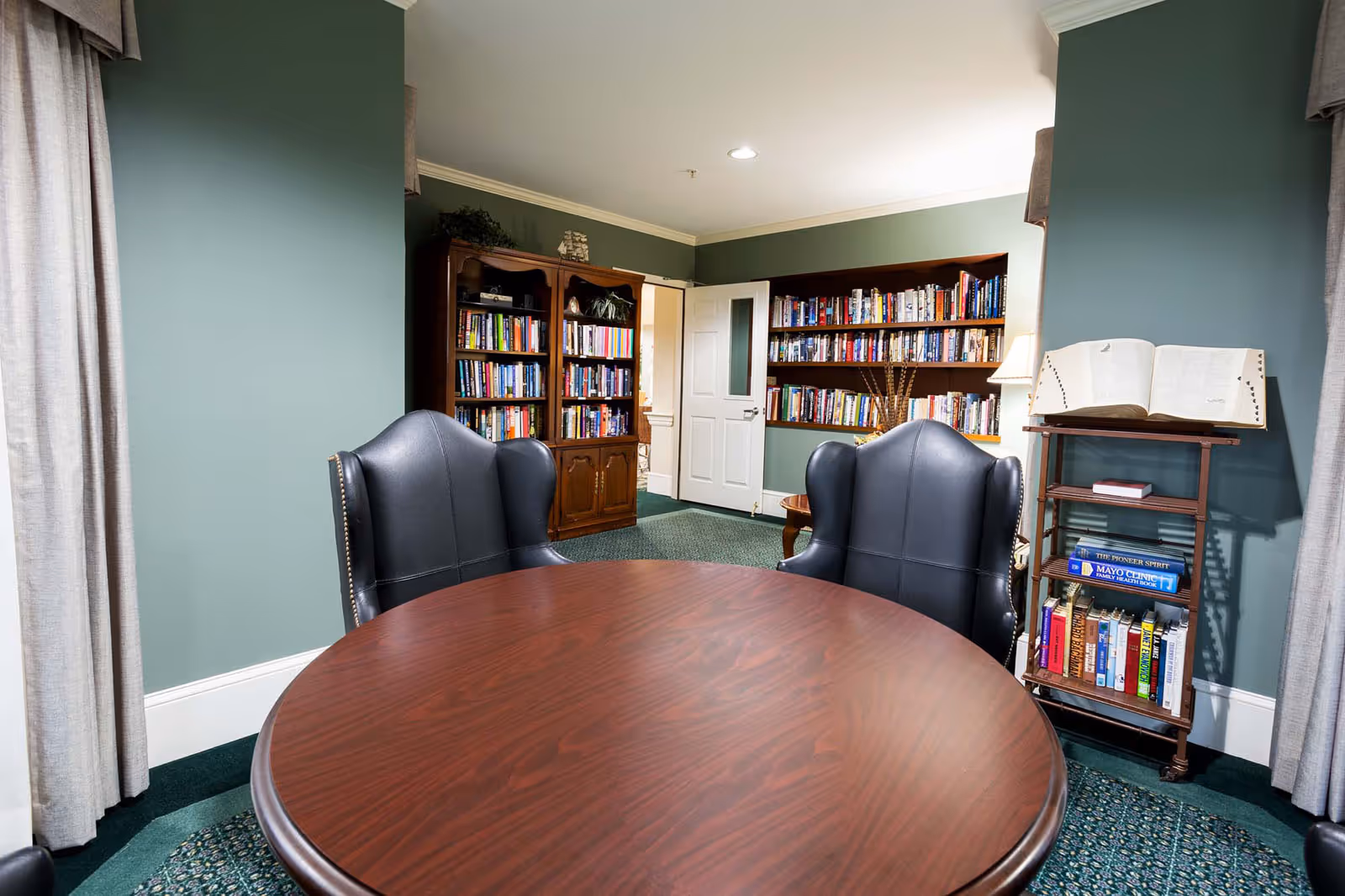 Round wooden table with two black leather chairs in a small library/reading room with bookshelves against green walls.