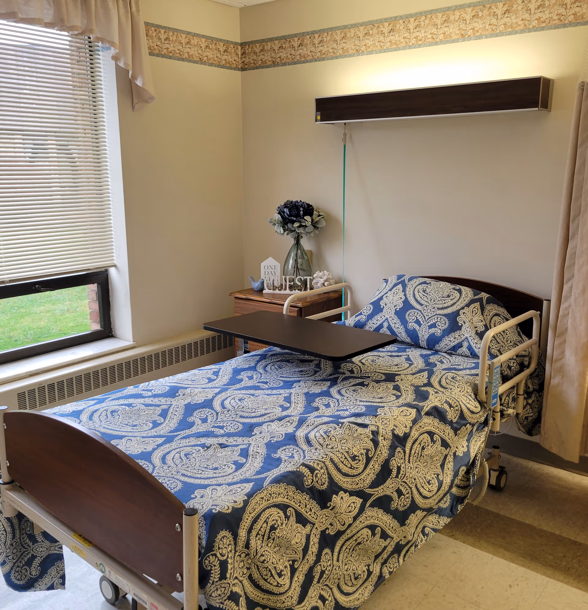 A nursing home bedroom with a hospital bed covered in a blue and beige patterned bedspread. The bed has side rails and an overbed table. Next to the bed is a wooden nightstand with a vase of flowers and decorative items. A window with blinds and a valance is on the left wall, and the room has beige walls with a floral border near the ceiling.