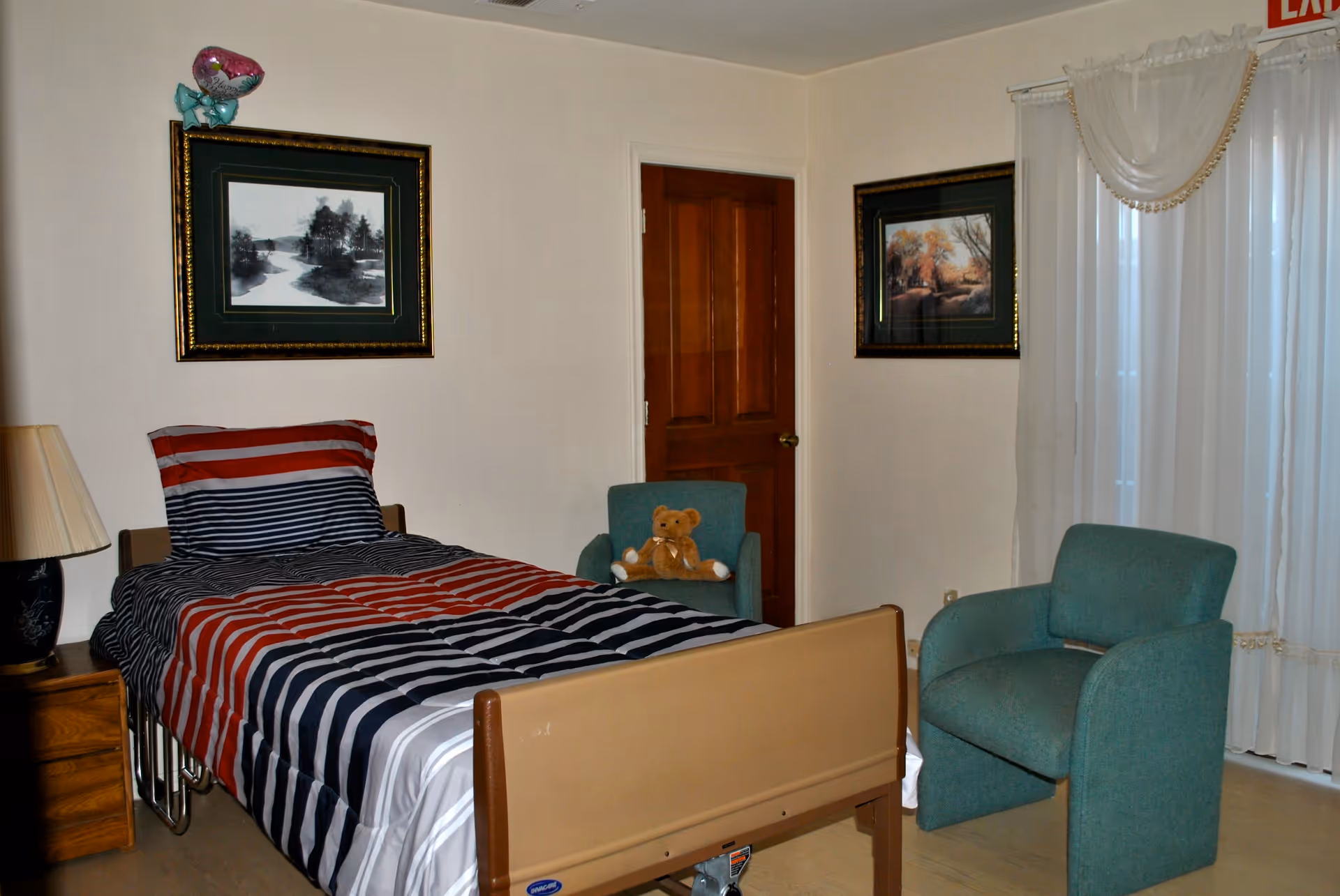 A bedroom in a senior living facility with a single bed covered in a striped red, white, and blue blanket. There is a wooden nightstand with a lamp on the left side of the bed. Two framed landscape paintings hang on the walls. A green armchair with a teddy bear sits near a closed wooden door, and another green armchair is positioned near a window with sheer white curtains. An exit sign is visible above the window.