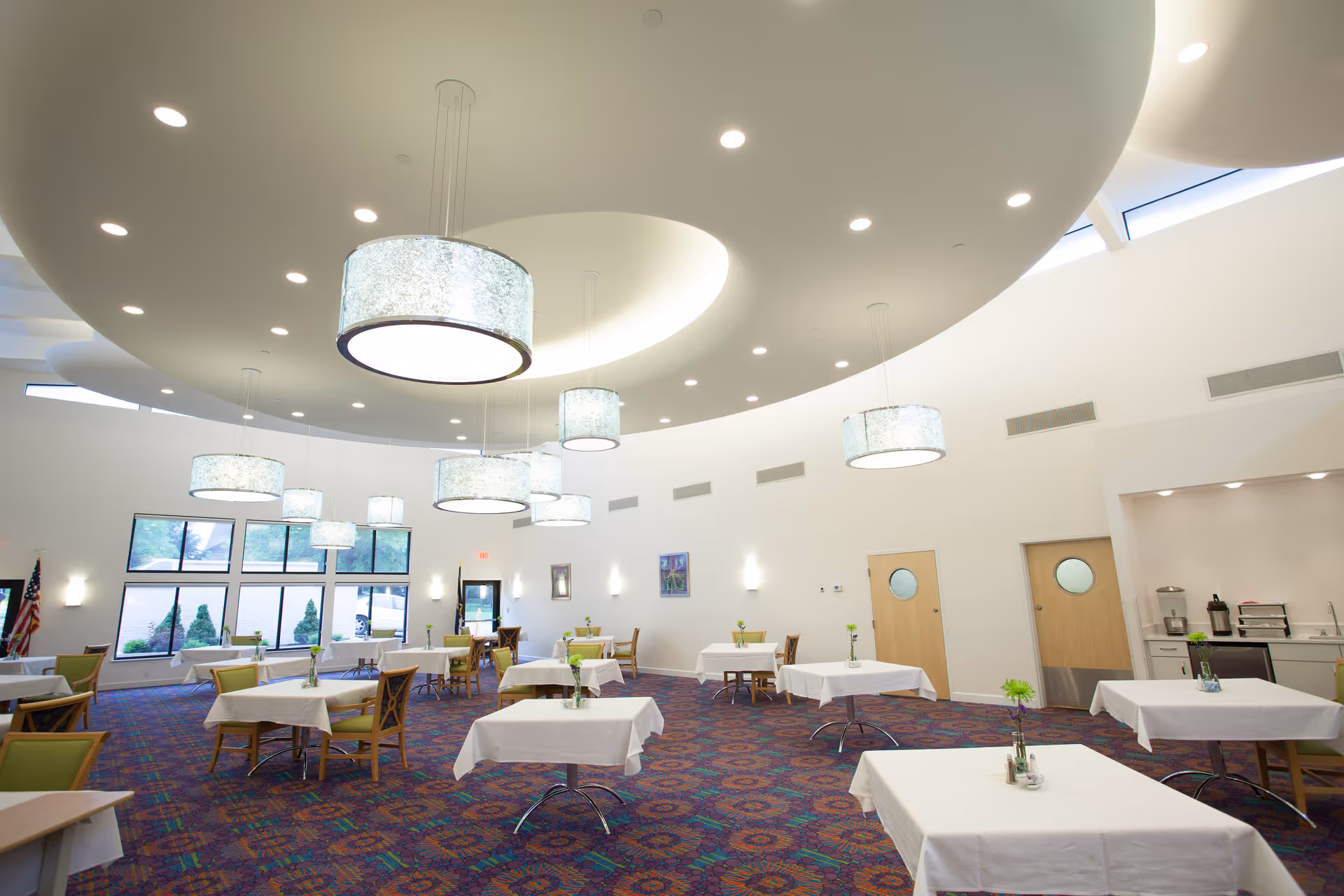 A spacious dining room with multiple tables covered in white tablecloths, each adorned with a small flower vase. The room features large circular ceiling lights, a patterned carpet, and large windows letting in natural light. There are wooden chairs with green cushions around the tables and a small kitchenette area in the back right corner.