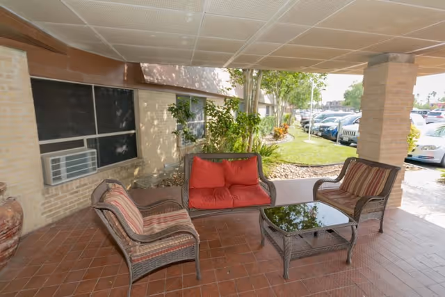 Covered outdoor seating area with wicker furniture including two armchairs with striped cushions, a loveseat with red cushions, and a glass-top coffee table. The area is paved with reddish tiles and supported by brick columns. There are plants and a grassy area visible beyond the seating area, along with parked cars in the background.