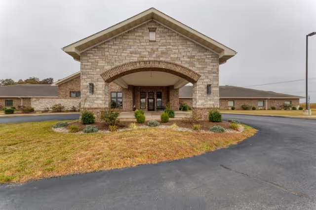 Front exterior view of a single-story senior living facility building with a stone and brick facade, a covered entrance with an arch, and a circular driveway surrounded by grass and small shrubs under an overcast sky.