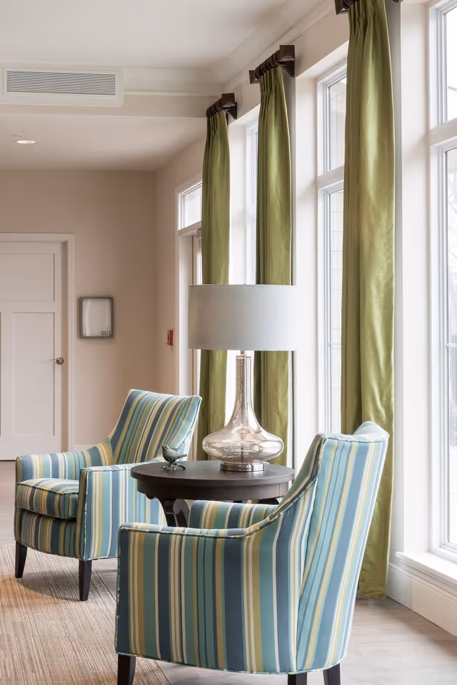 A cozy sitting area with two striped armchairs in shades of blue, green, and beige positioned around a round wooden table with a decorative glass lamp and a small bird figurine. The space is well-lit by large windows with green curtains.