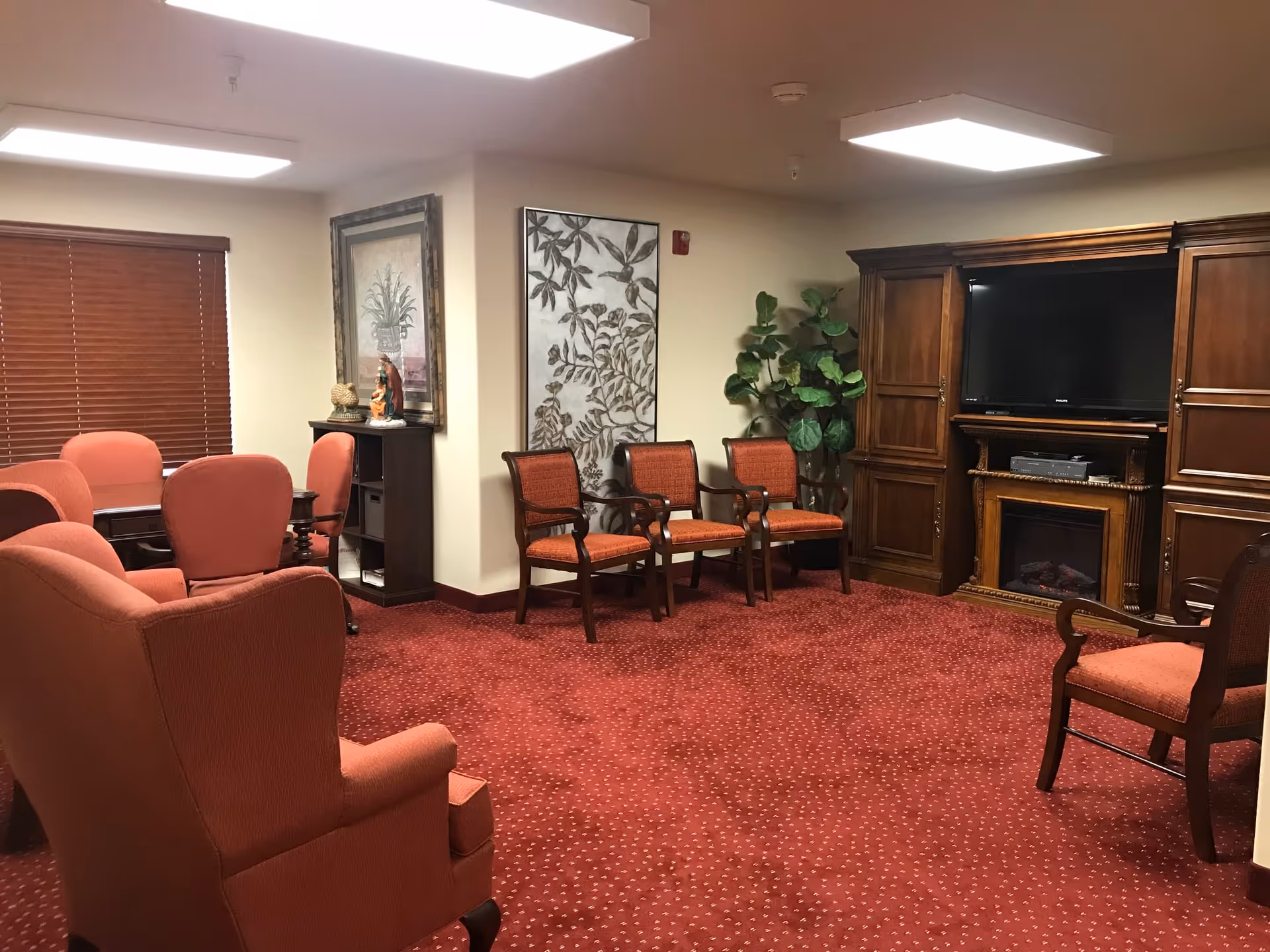A cozy senior living common area with red carpet and matching upholstered chairs arranged around the room. There is a large wooden entertainment center with a flat-screen TV and an electric fireplace. The walls are decorated with framed artwork and a large plant is placed in the corner. The room is lit by ceiling lights and has a window with wooden blinds.