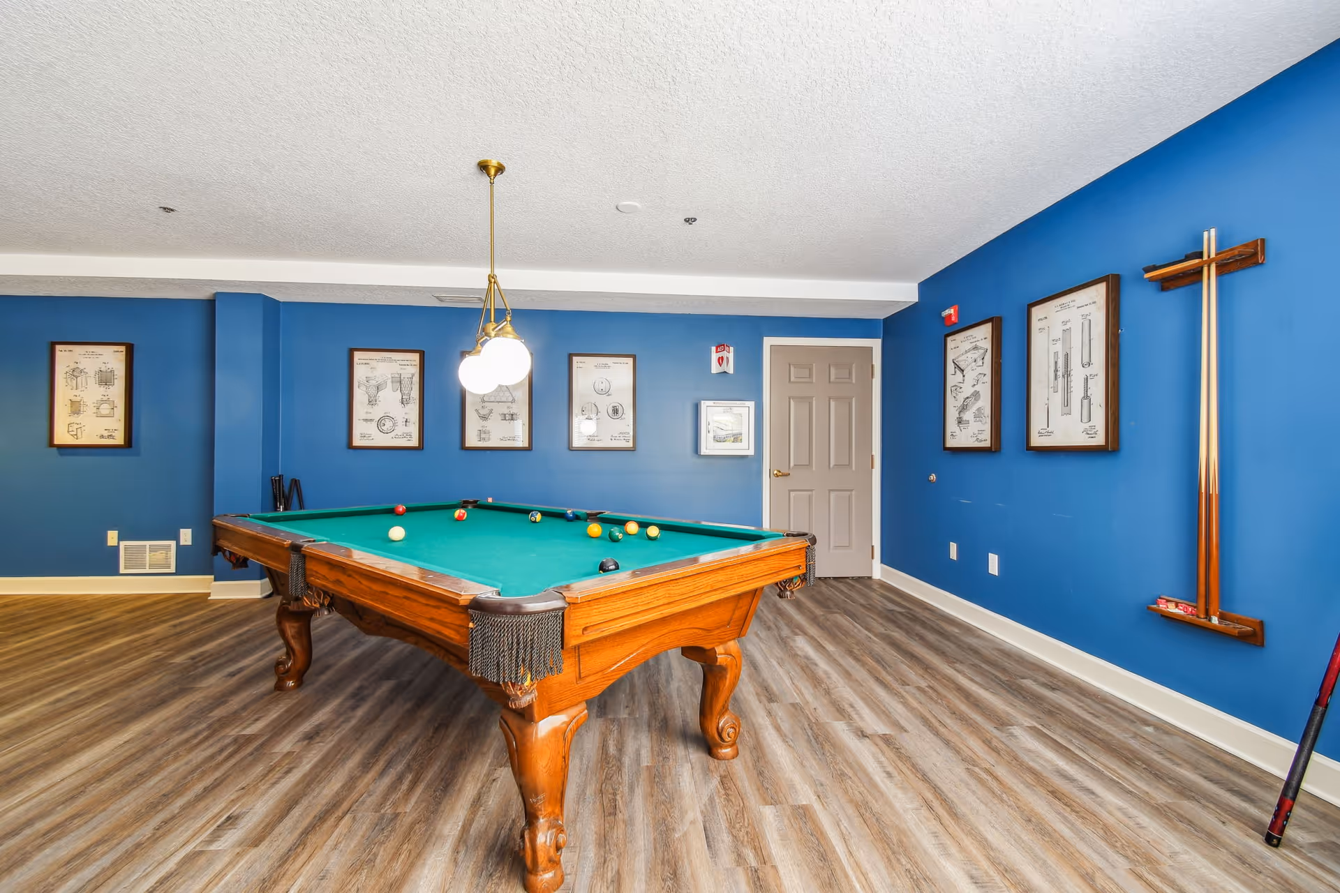 A game room with a wooden pool table featuring a green felt surface and billiard balls arranged on it. The room has blue walls adorned with framed vintage-style prints and a rack holding pool cues. The floor is covered with wood-patterned vinyl or laminate flooring, and a hanging light fixture with three bulbs is positioned above the pool table.
