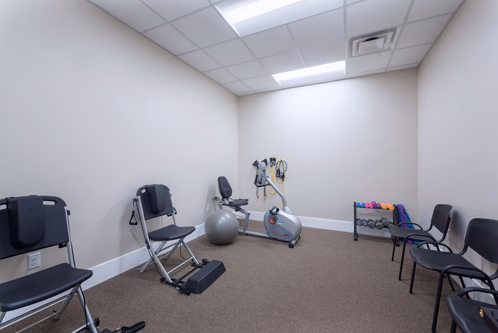 A small exercise room with beige walls and carpeted floor, containing two black exercise chairs, a recumbent exercise bike, an exercise ball, resistance bands hanging on the wall, a rack with various dumbbells and yoga mats, and three black chairs lined up against the right wall.