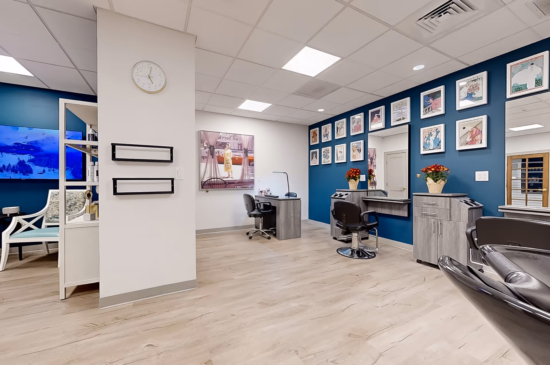 Interior view of a salon area in a senior living facility with blue accent walls adorned with framed artwork, salon chairs, a manicure table, a clock on a white pillar, and a television in the adjacent room.