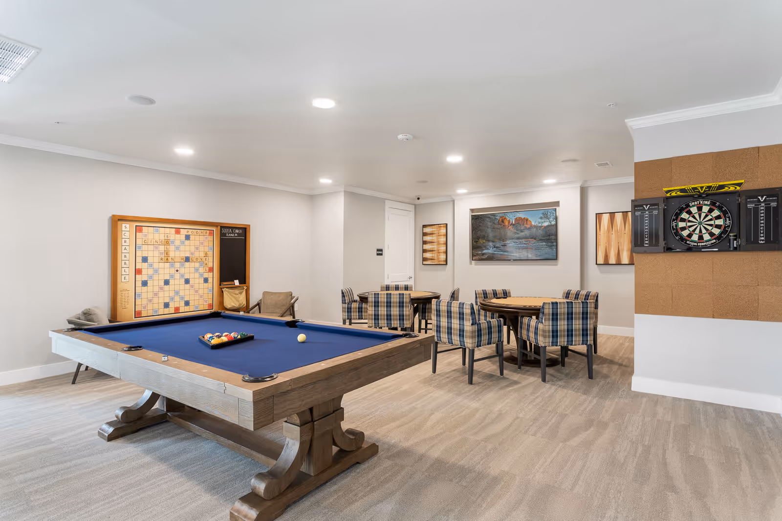 A recreational room with a blue felt pool table in the foreground, a large Scrabble board on the wall, two round tables with plaid upholstered chairs, a dartboard mounted on a corkboard wall, and framed artwork on the walls.