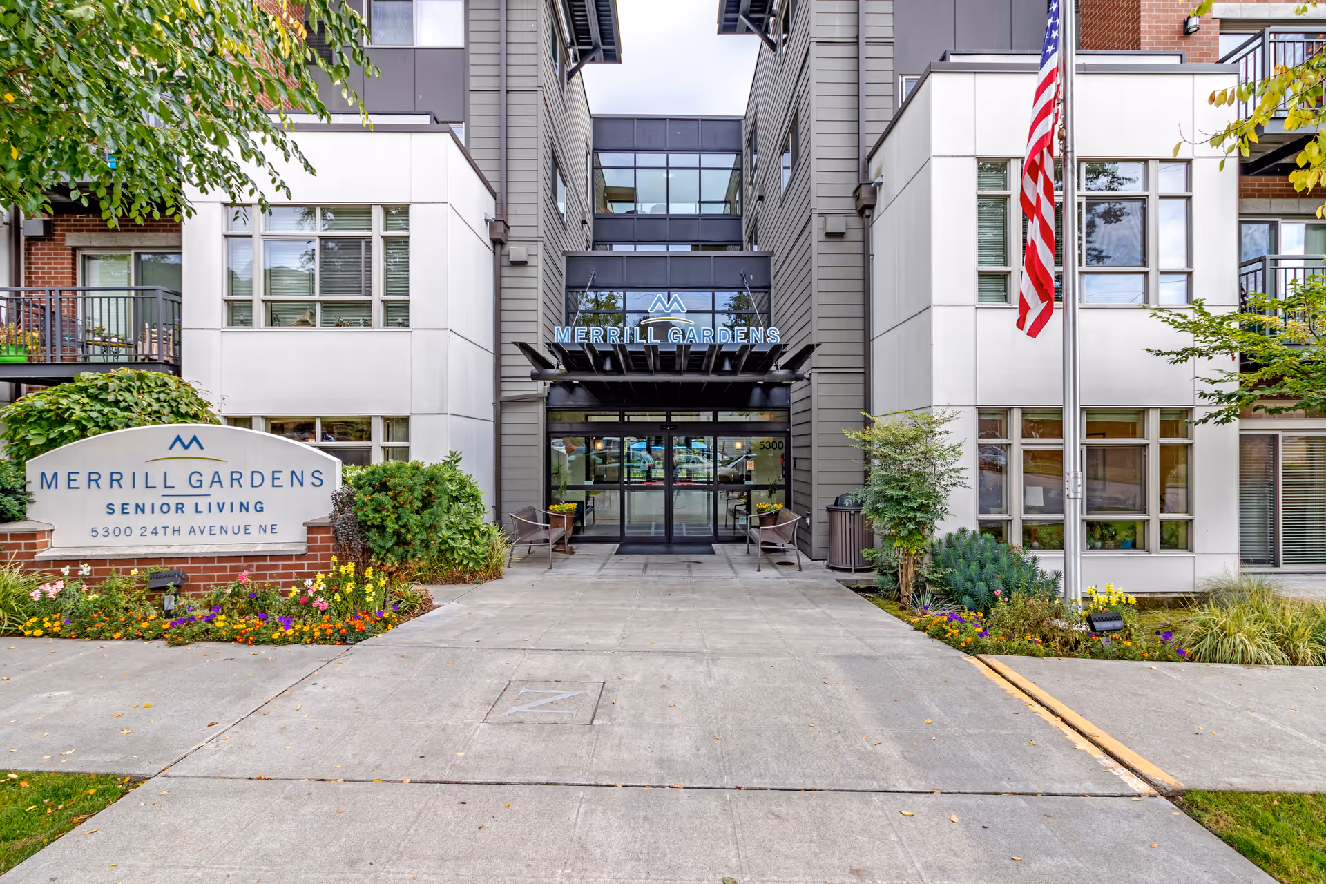 Front exterior view of Merrill Gardens Senior Living facility with a clear entrance, American flag on a pole, and a sign displaying the facility name and address surrounded by flowers and greenery.