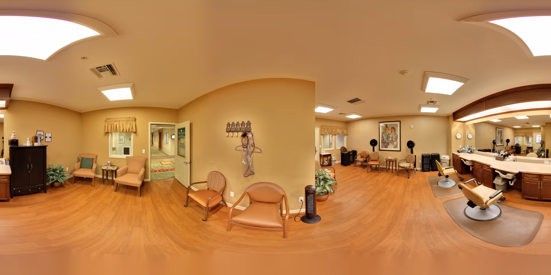 A spacious and well-lit salon area in a senior living facility with wooden flooring, beige walls, and ceiling lights. The room features two salon chairs in front of a large mirror with a counter holding various hair care products. There are several comfortable chairs arranged along the walls, a small table with a plant, and a coat rack with a hairdryer hanging on it. A hallway is visible through an open door, and framed artwork decorates the walls.