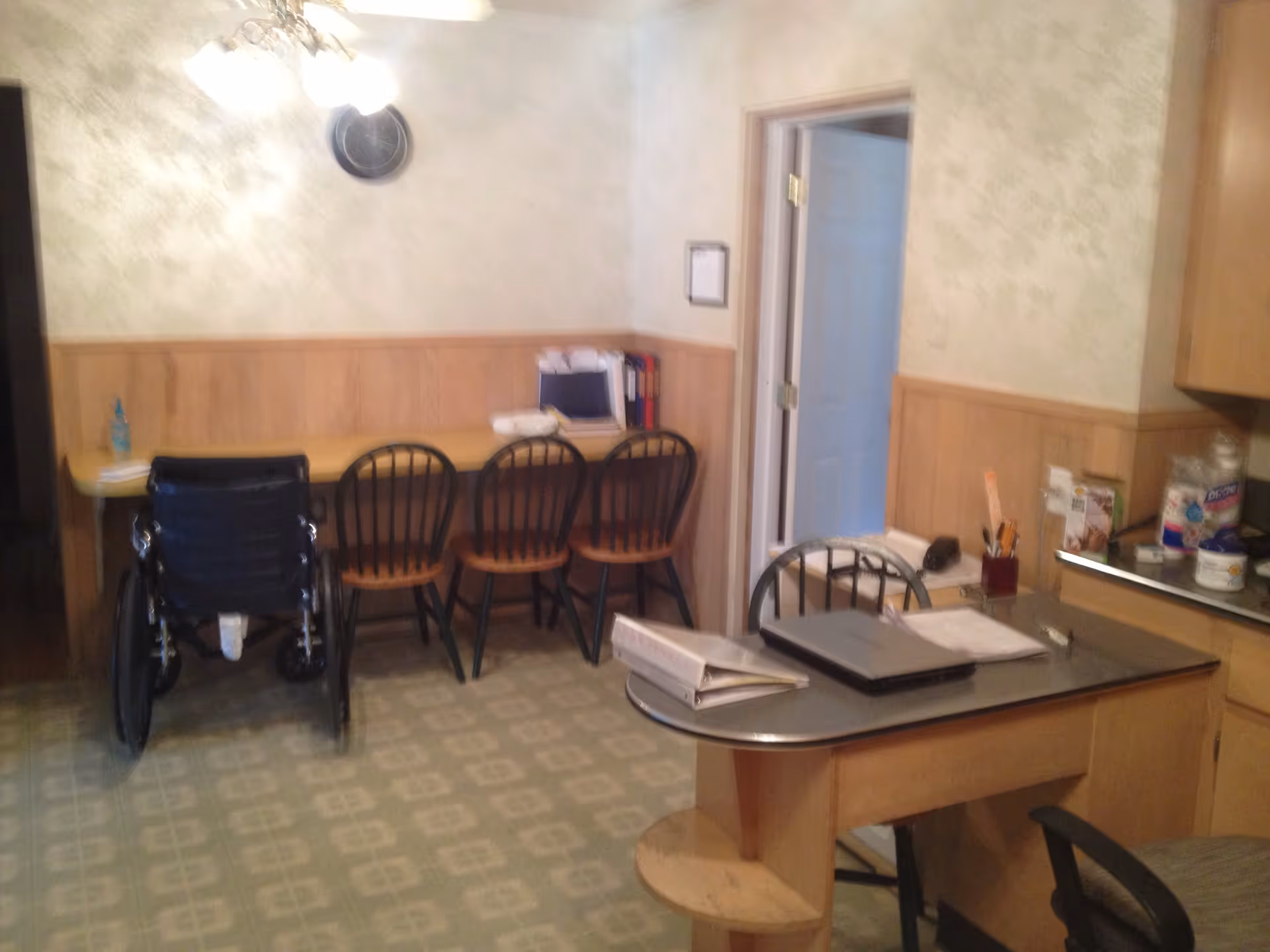 Interior room with a long counter against the wall with four chairs, including a wheelchair, and a small table with a laptop, binder, and office supplies. The walls have wood paneling and light-colored wallpaper, and there is a door leading to another room.