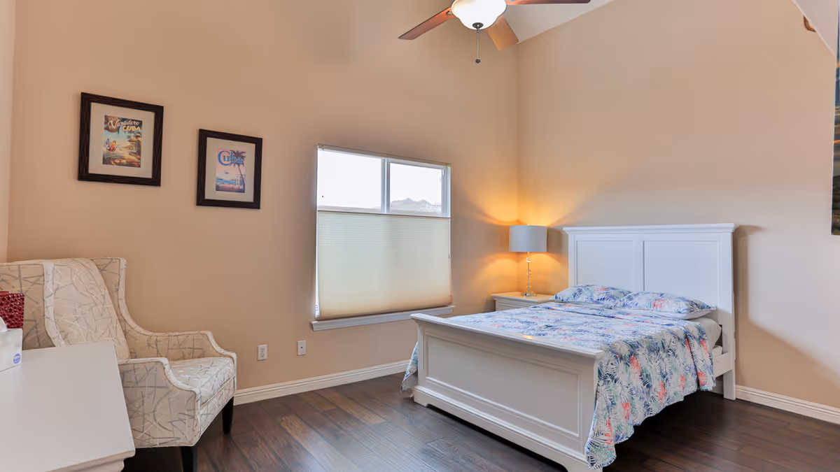 A bright and clean bedroom with a white wooden bed frame and a colorful floral bedspread. There is a white nightstand with a lamp emitting warm light next to the bed. A window with a partially closed blind is on the wall behind the bed. On the left side, there is a patterned armchair and two framed pictures hanging on the beige wall. The room has wooden flooring and a ceiling fan with a light fixture.