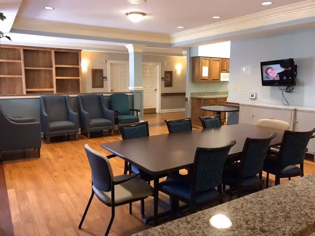A spacious common area in a senior living facility featuring a large rectangular table surrounded by chairs in the foreground. In the background, there are several armchairs arranged along the wall, wooden shelves, and a small kitchen area with wooden cabinets. A flat-screen TV is mounted on the wall showing a person on screen. The room has wooden flooring and soft lighting from ceiling fixtures.