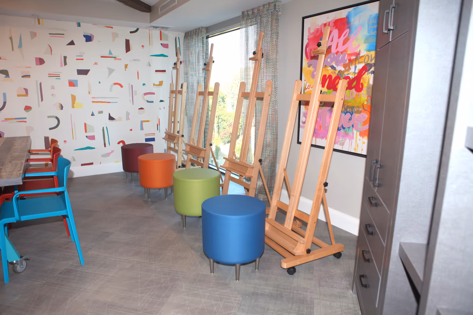 A bright art room with colorful stools in blue, green, orange, and maroon lined up in front of wooden easels. The room has a large window with patterned curtains, a wall with abstract colorful shapes, and a vibrant framed artwork with words like 'Love' and 'All'. There are also blue and red chairs around a wooden table on the left side and gray cabinets on the right.