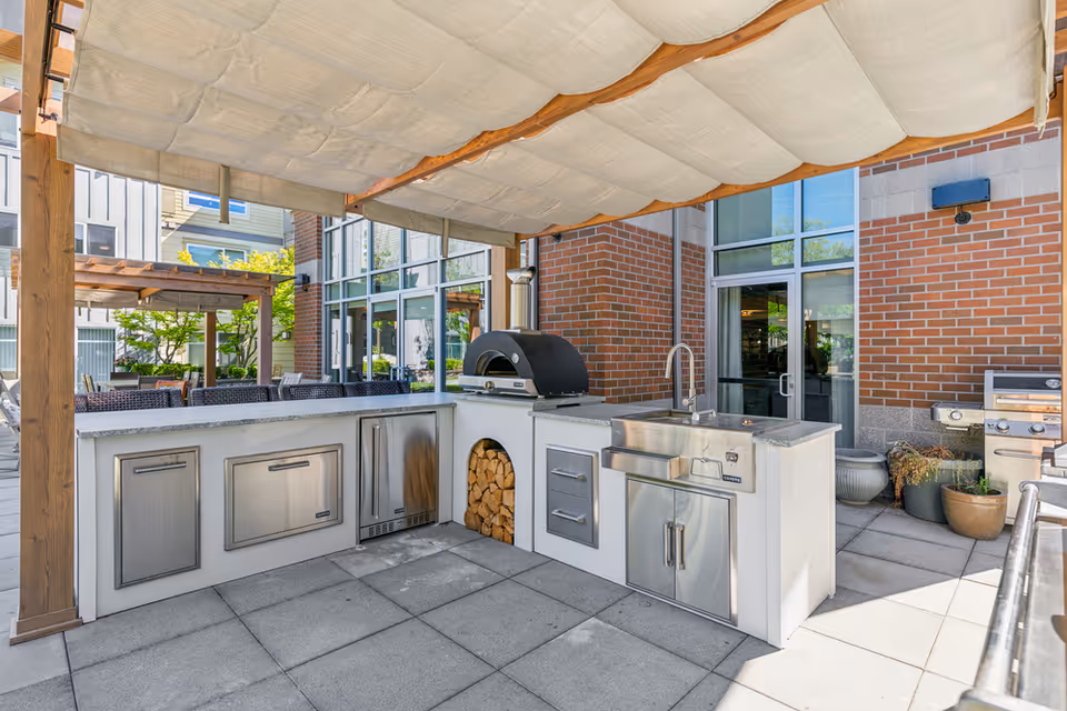 Outdoor kitchen area with stainless steel appliances including a grill, sink, and storage cabinets under a fabric canopy. The area is paved with large tiles and surrounded by brick walls and glass doors, with some potted plants nearby.