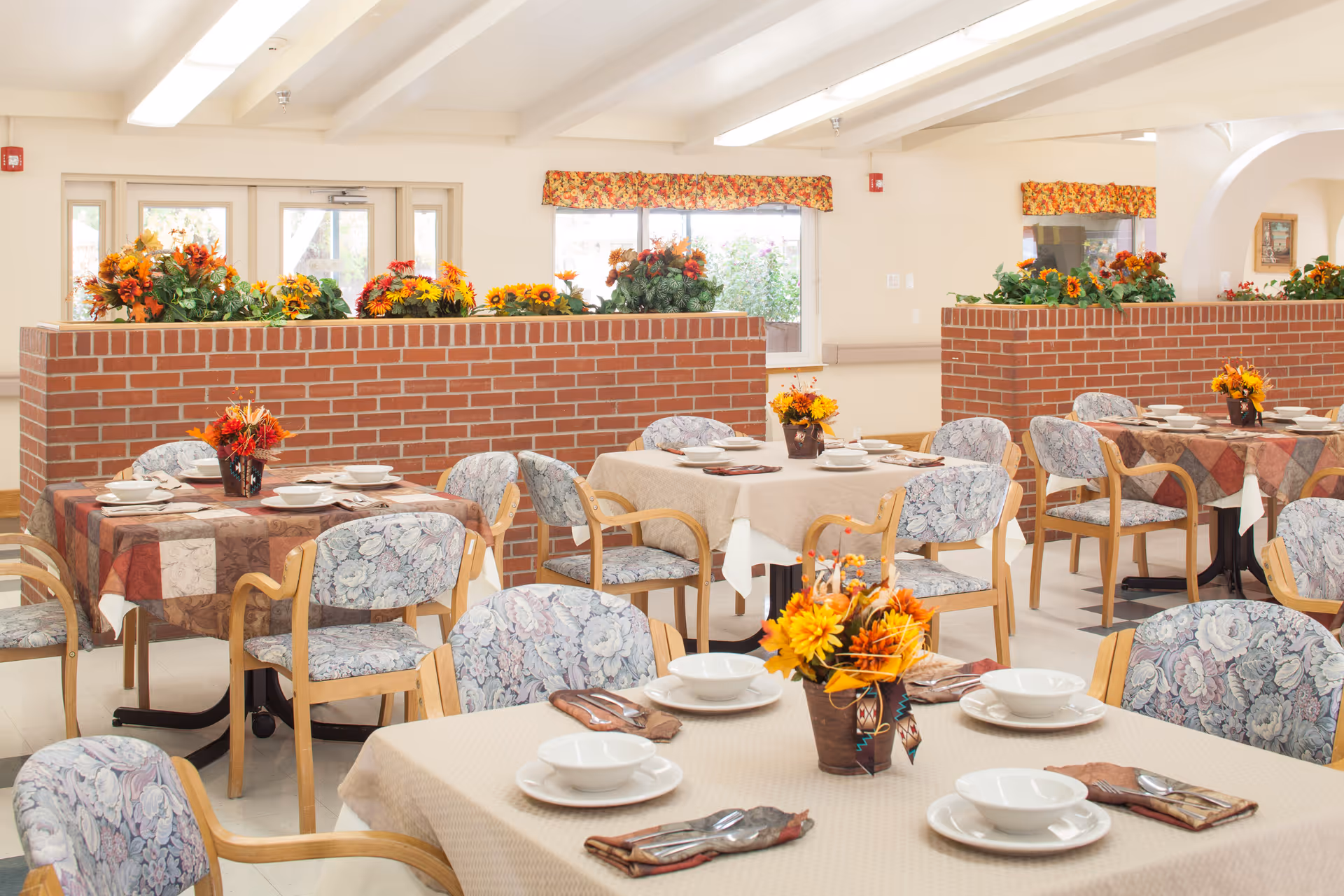 A bright dining room with multiple tables covered in beige and patterned tablecloths, each set with white bowls, plates, and silverware. The chairs have wooden frames and floral upholstery. There are brick half-walls decorated with vibrant orange and yellow flower arrangements, and windows with floral valances letting in natural light.