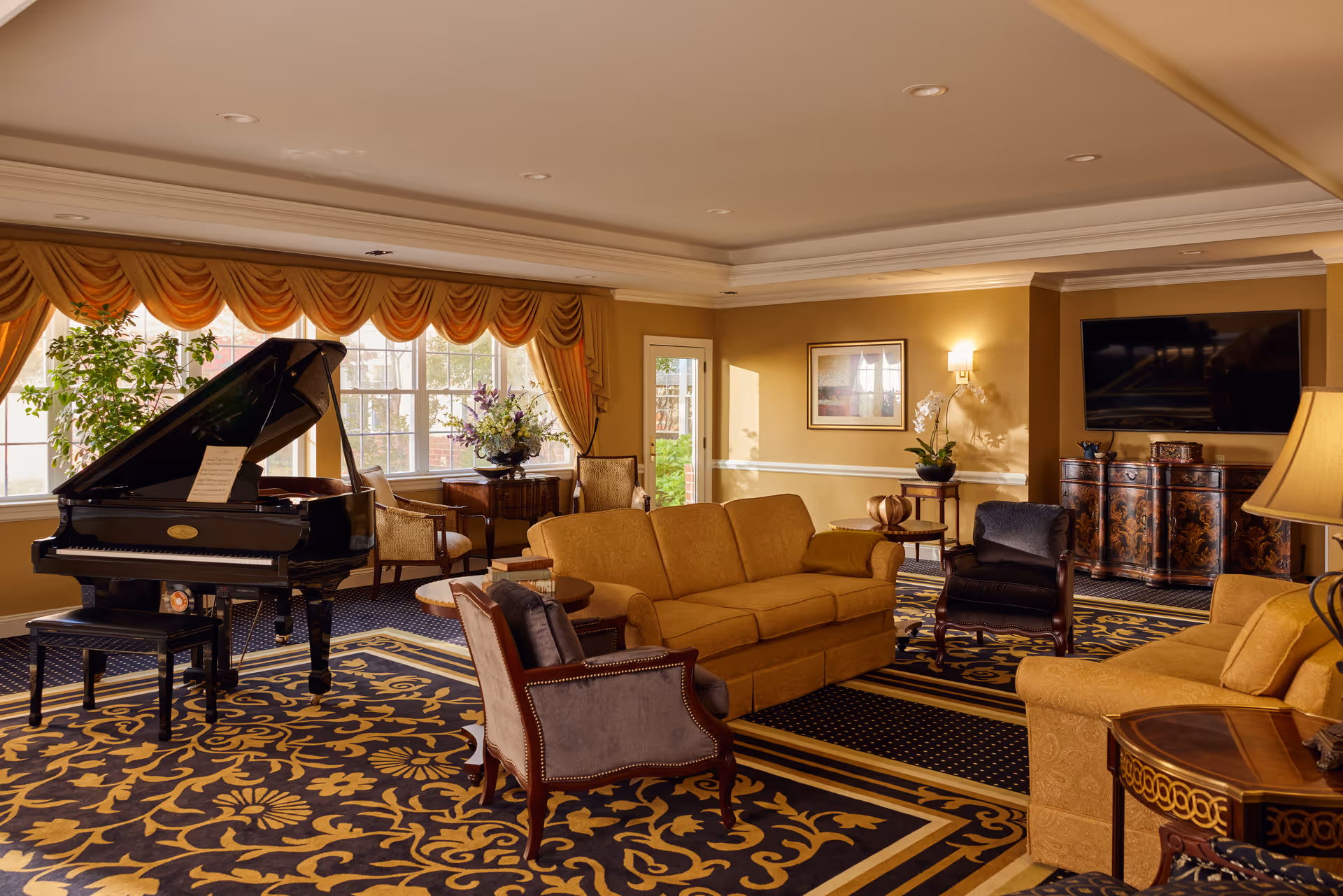 A spacious and elegant living room with a black grand piano on the left side near large windows with gold drapes. The room features two mustard yellow sofas, two gray armchairs, a patterned carpet with gold and navy blue designs, a flat-screen TV mounted on the wall, and several decorative tables with plants and flowers. Warm lighting enhances the cozy and inviting atmosphere.