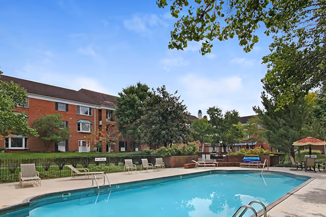 Outdoor swimming pool with lounge chairs and umbrellas in a courtyard in front of a brick senior living building under a blue sky.
