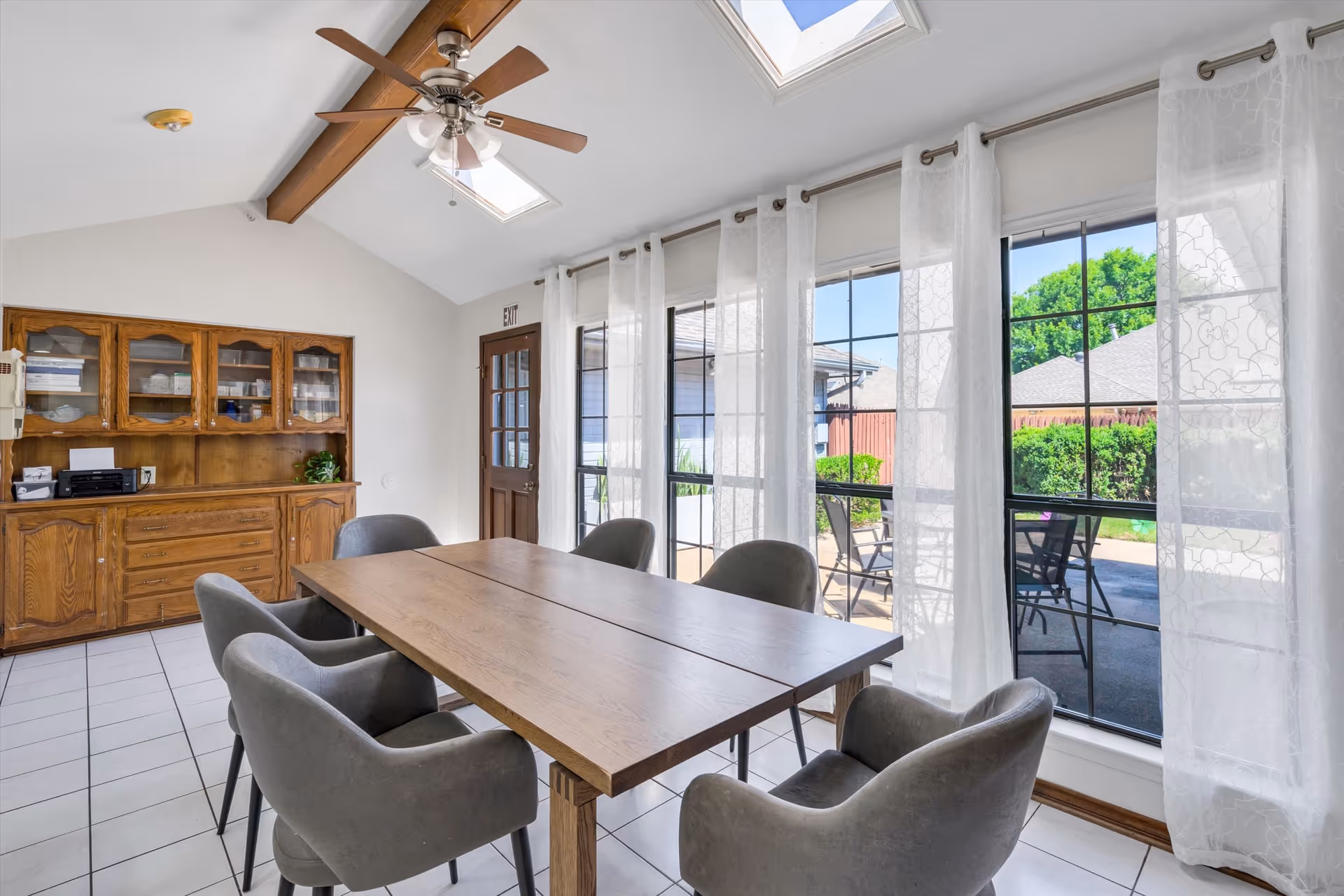 Bright dining room with a wooden table surrounded by gray upholstered chairs, large windows with sheer curtains, skylights, and a wooden hutch.