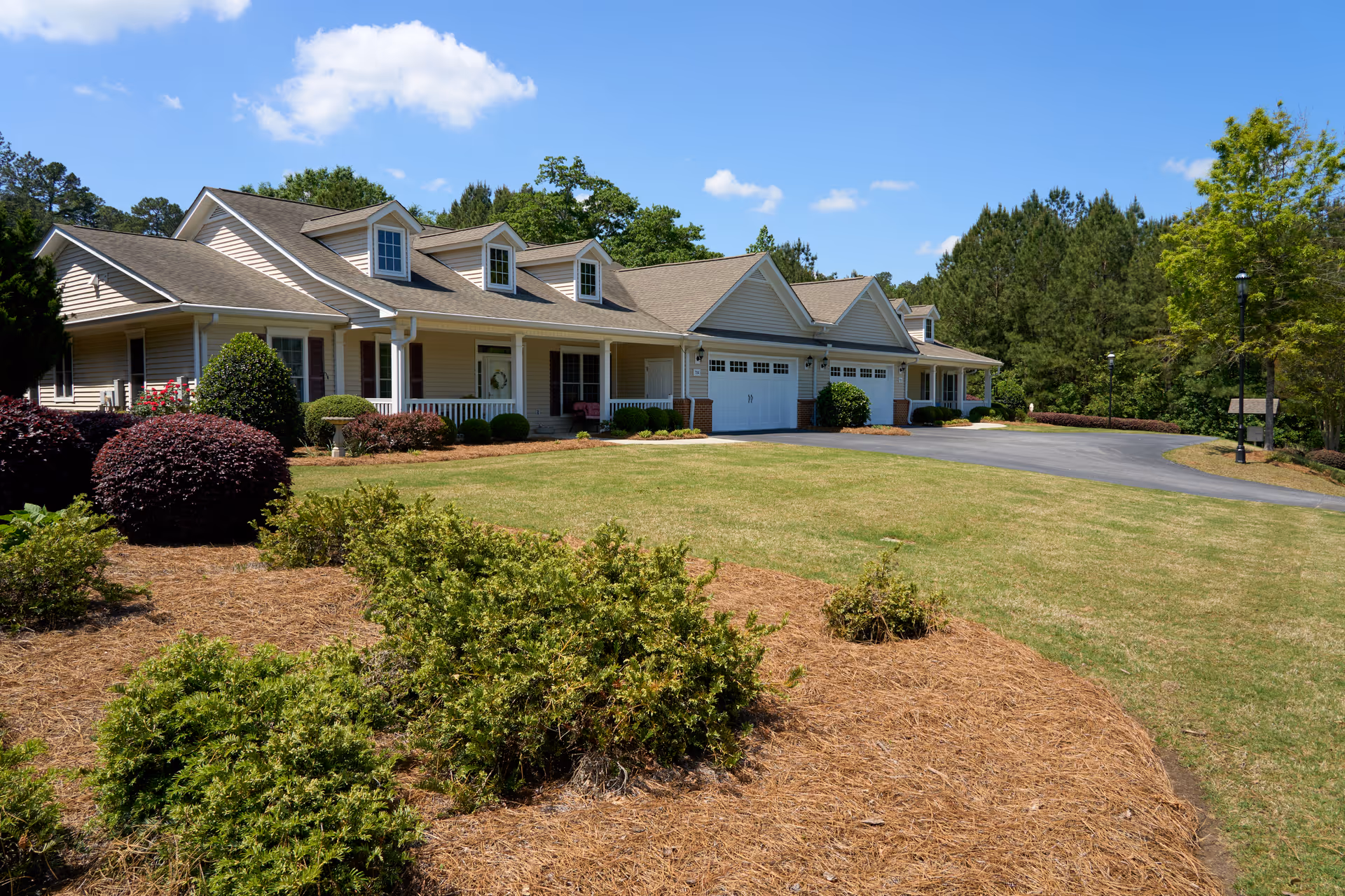 Exterior view of a single-story residential building with beige siding, multiple dormer windows, a covered front porch, and attached garages. The building is surrounded by a well-maintained lawn, landscaped bushes, and trees under a blue sky with some clouds.