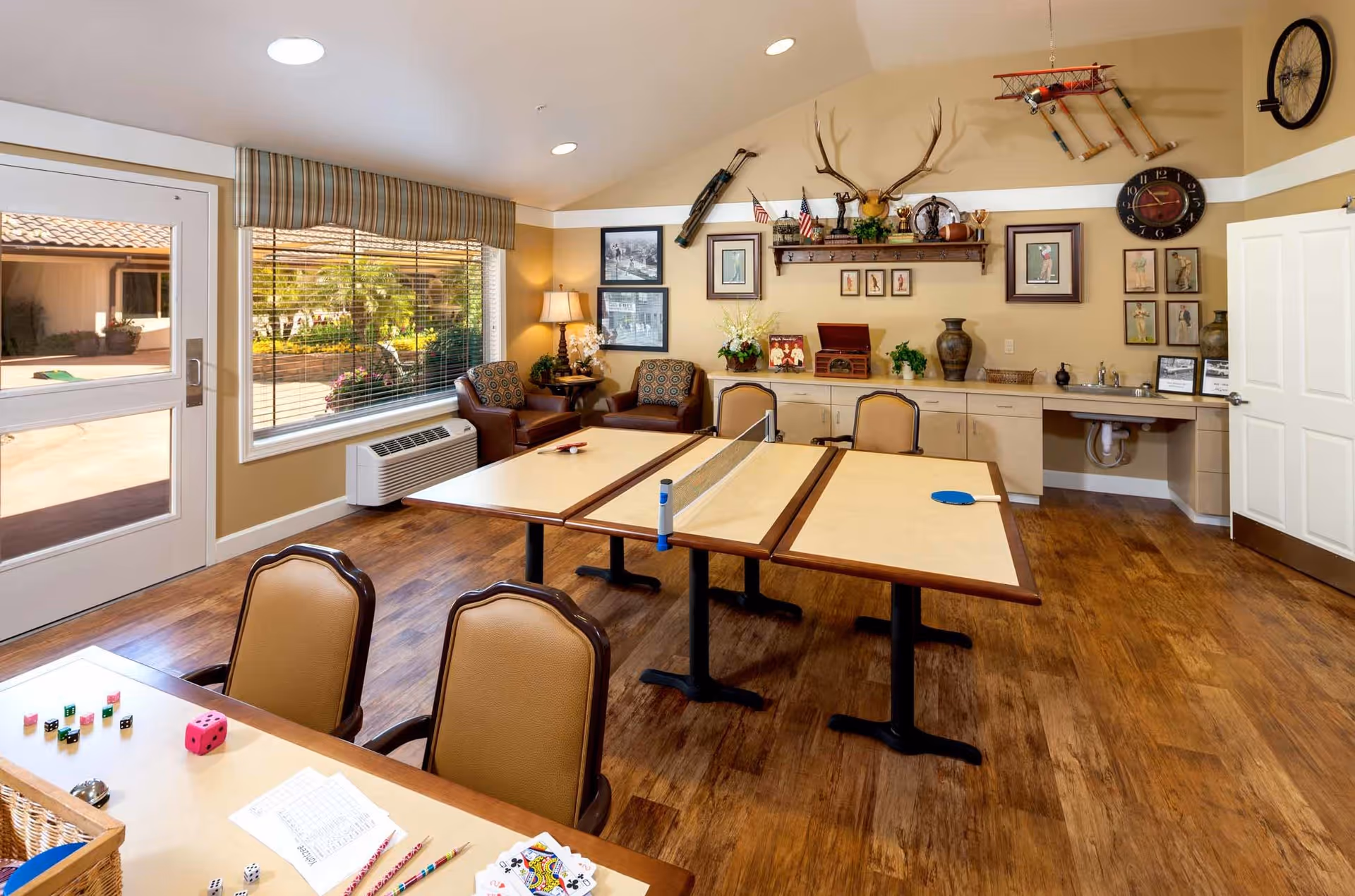 A bright and cozy activity room with a ping pong table in the center, two cushioned chairs with patterned pillows near a window, a table with playing cards, dice, and pencils, and a wall decorated with framed pictures, a mounted deer head, a model airplane, and a clock. There is a door leading outside and a countertop with cabinets and a small sink.