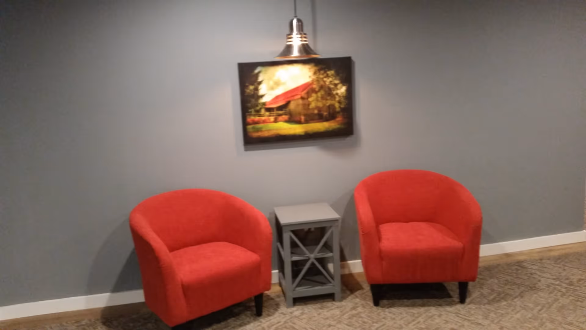 Two red upholstered armchairs with black legs are positioned on either side of a small gray side table with a crisscross design. Above the table hangs a framed picture of a rustic barn with a red roof, illuminated by a hanging light fixture. The setting is against a gray wall with a beige carpeted floor.