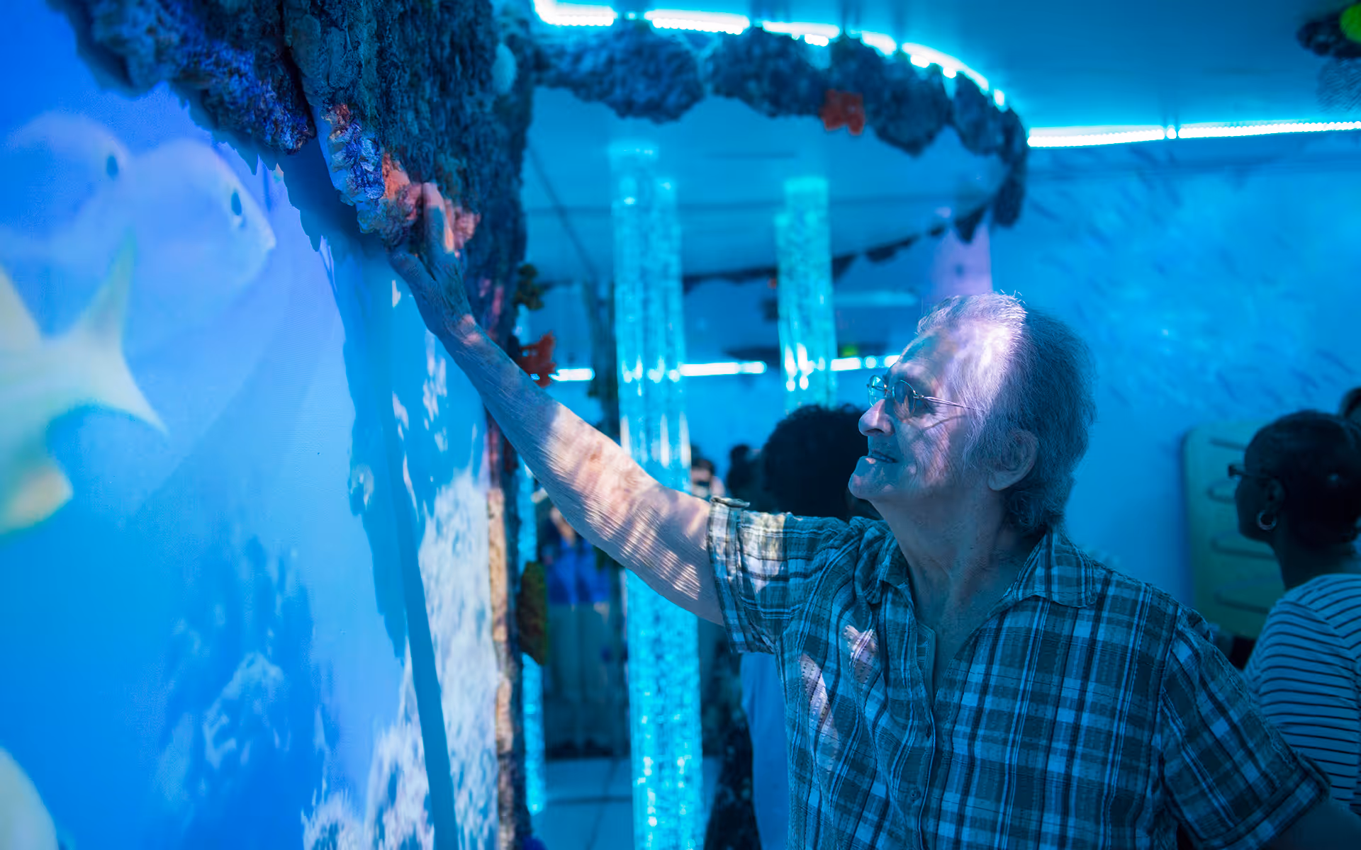 An elderly woman wearing glasses and a plaid shirt reaches out to touch a textured wall with an underwater theme, illuminated by blue lighting. Other people are visible in the background, also engaged with the exhibit.
