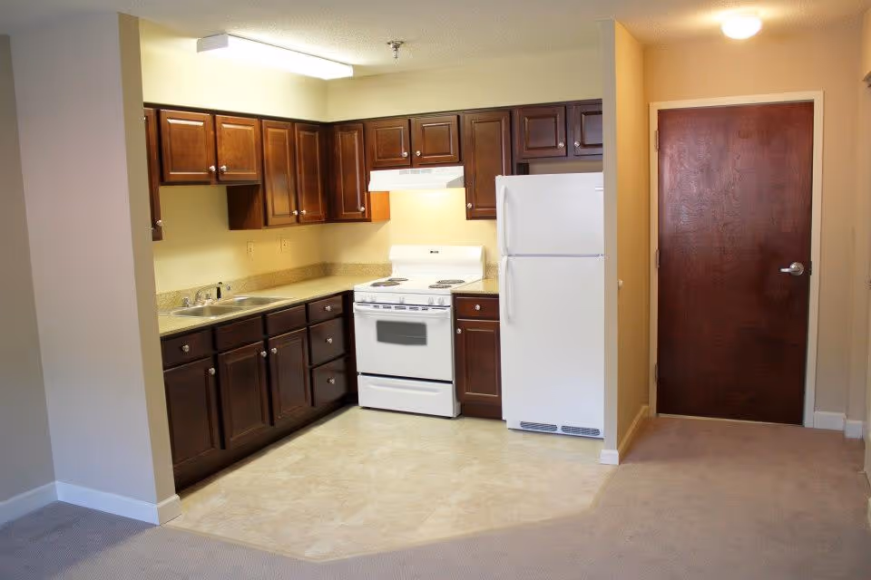 Interior view of a kitchen area in a senior living facility with dark wood cabinets, a white stove, white refrigerator, beige countertops, and beige tiled flooring. Adjacent to the kitchen is a carpeted area and a closed wooden door with a silver handle.