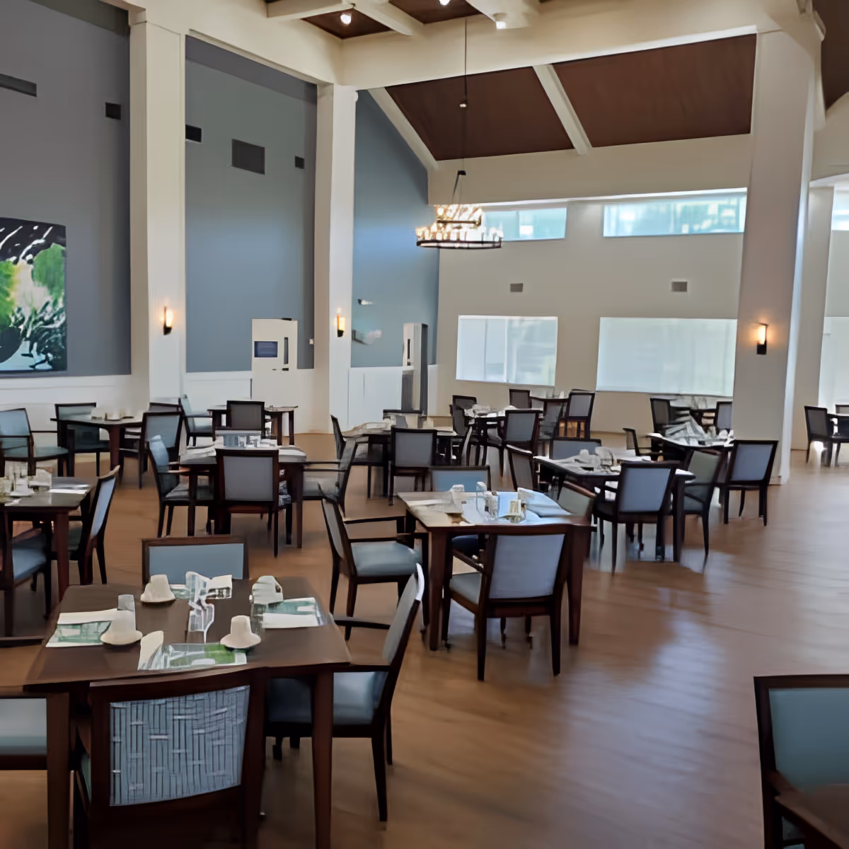 A spacious dining room with multiple wooden tables and chairs arranged neatly. Each table is set with placemats, napkins, and glassware. The room features high ceilings with exposed beams, large windows allowing natural light, and modern wall sconces providing additional lighting. A large abstract painting is visible on one wall.