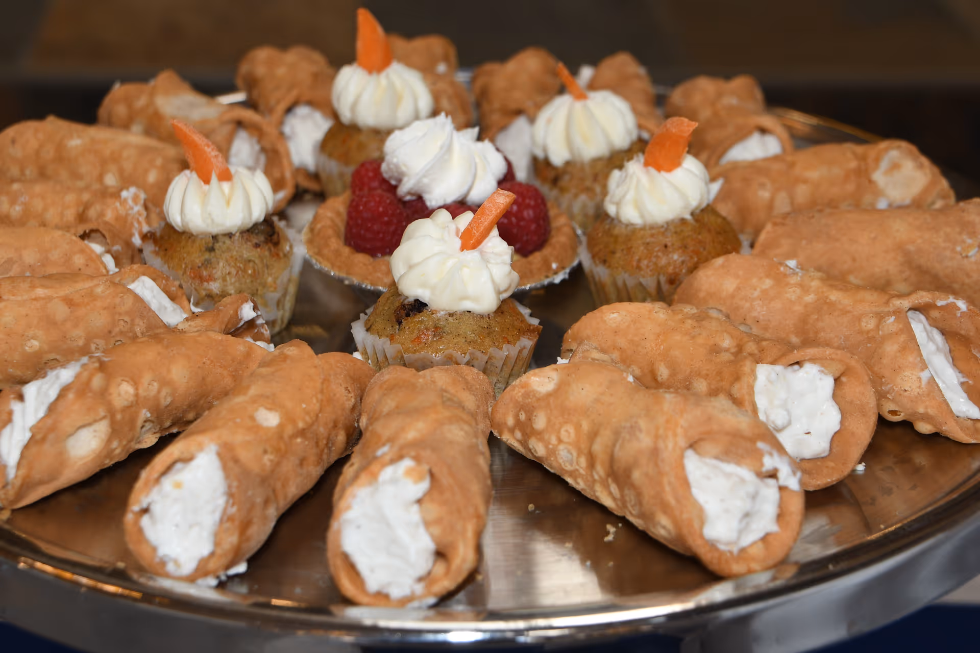 A close-up view of a silver tray filled with cannoli pastries and small cupcakes topped with whipped cream and a small piece of orange garnish, along with raspberries on some cupcakes.