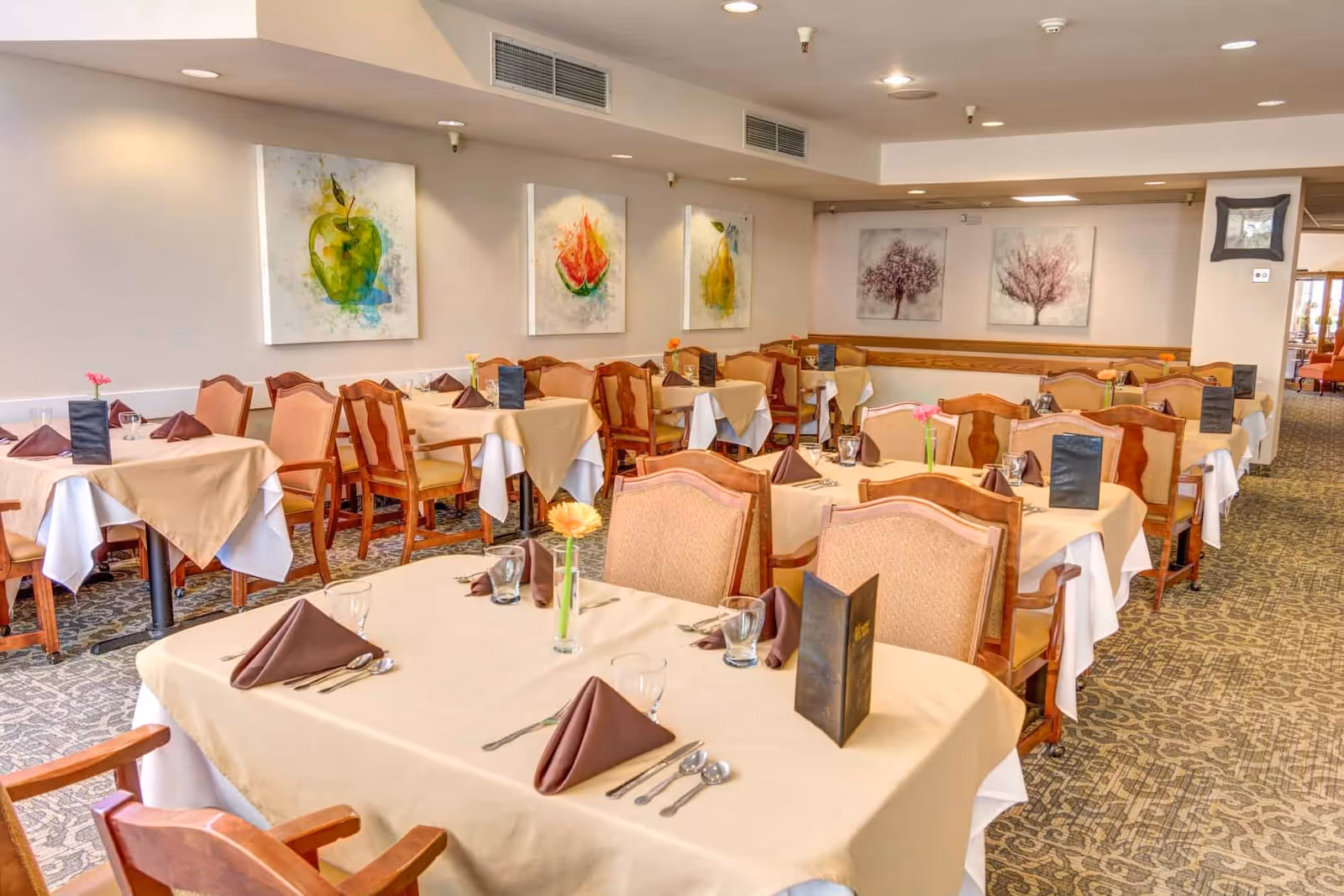 A dining room with multiple tables covered in beige and white tablecloths, each set with brown folded napkins, glassware, cutlery, and small flower vases. The room has wooden chairs with cushioned seats and backs. The walls are decorated with colorful paintings of fruit and trees. The carpet has a patterned design, and the room is well-lit with recessed ceiling lights.