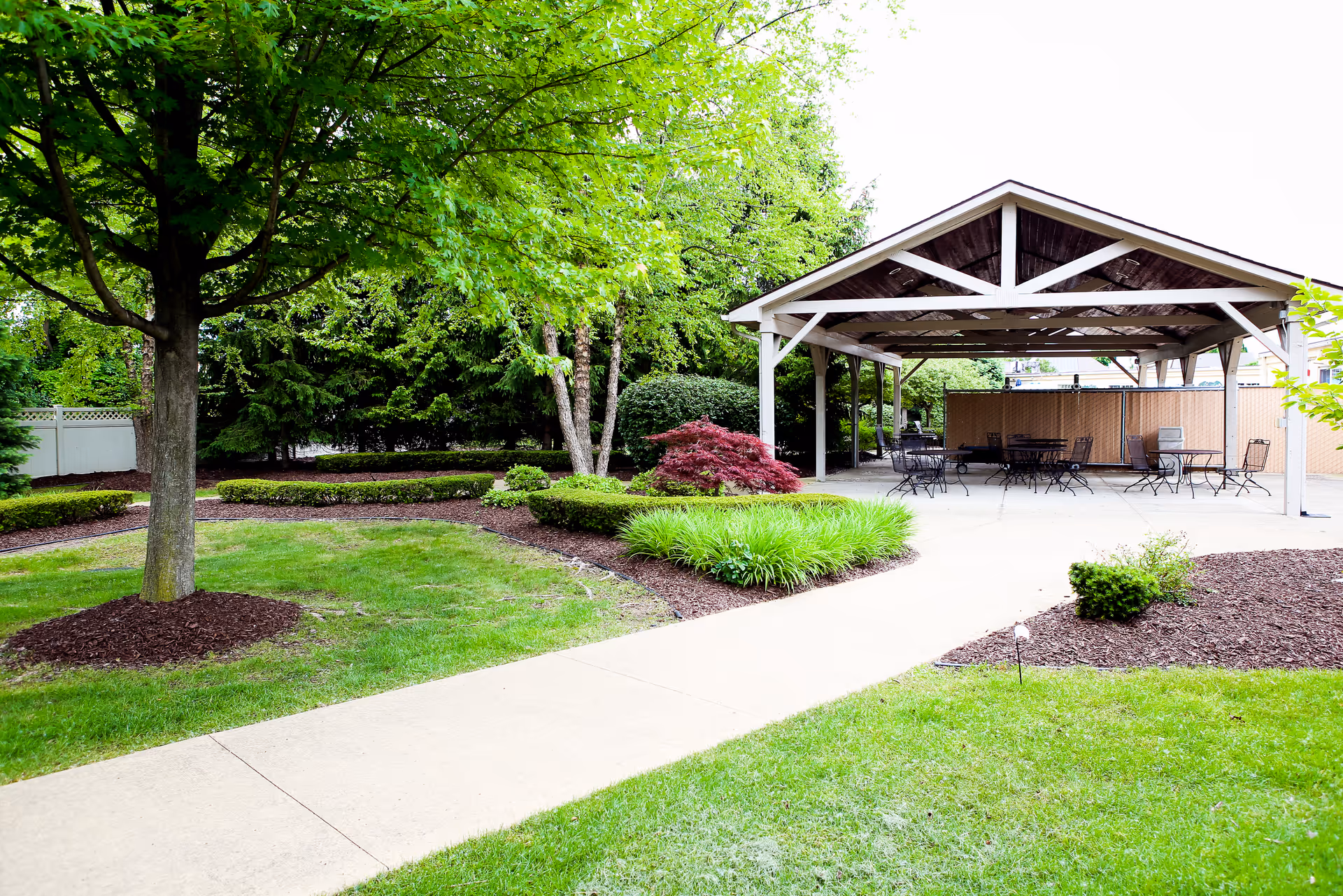 Outdoor area with a paved walkway leading to a covered patio with several metal tables and chairs. The surrounding landscape includes green grass, mulched garden beds, bushes, and trees providing shade.