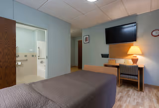 A room with a single bed covered in a brown quilt, a wooden desk with a lamp and a chair, a wall-mounted flat-screen TV, and an open door leading to a bathroom with grab bars and a bathtub.