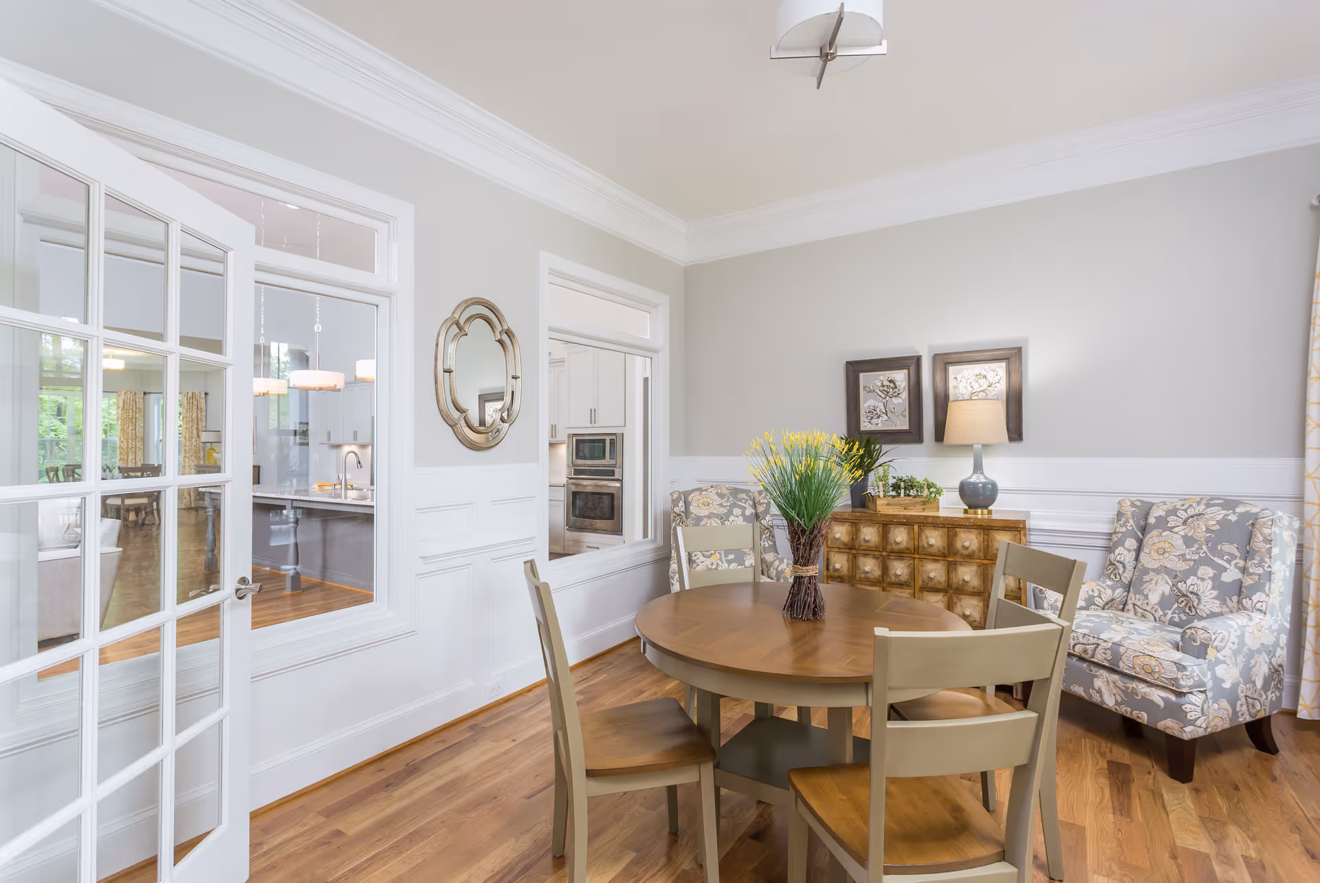A bright dining room with a round wooden table surrounded by four chairs. A floral-patterned armchair and a wooden sideboard with a lamp and framed artwork are against the wall. The room has light gray walls with white wainscoting and hardwood floors. A glass-paned door and window reveal a kitchen area in the background.