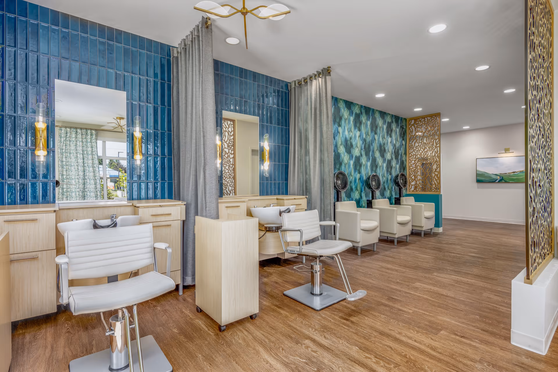 Interior view of a modern salon area with white styling chairs and wash basins in front of blue tiled walls with mirrors and wall lights. The floor is wooden, and there are three white hair dryer chairs against a patterned blue-green accent wall. Decorative gold panels partially separate the space.