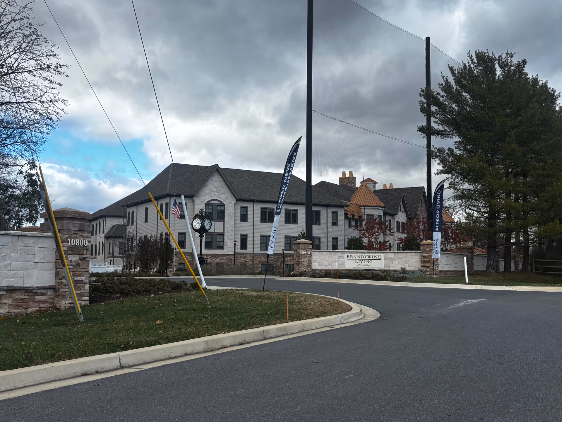 Exterior view of Brandywine Living At Potomac, a senior living facility with a large multi-story building, stone walls, and a clock near the entrance. The sky is cloudy with some patches of blue, and there are trees and flags near the entrance.