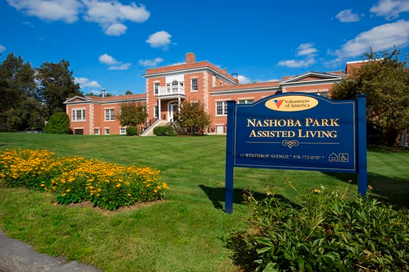 Exterior view of Nashoba Park Assisted Living, a large brick building with white trim, surrounded by green lawn and yellow flowers under a blue sky with some clouds. A blue sign in the foreground displays the facility name, address, phone number, and the Volunteers of America logo.