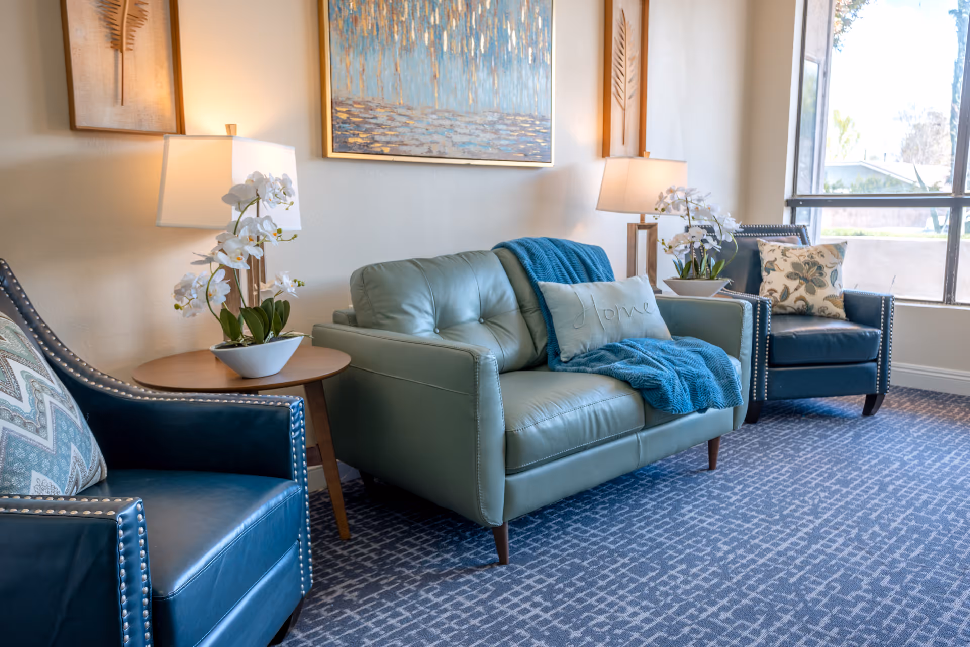 Bright seating area with a teal leather loveseat and matching armchairs, side tables with lamps and orchids, and a large window.