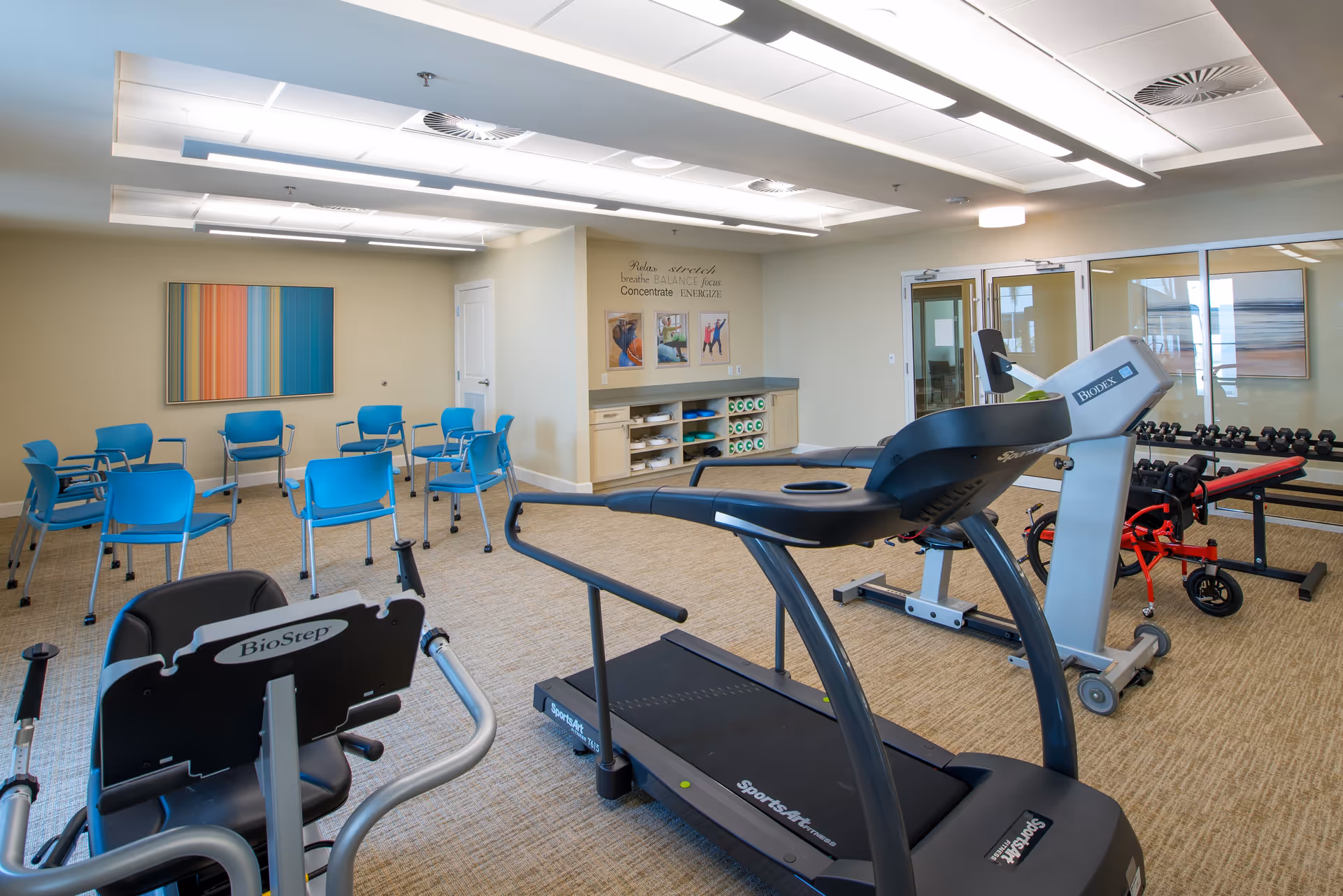 A fitness and exercise room with various equipment including a treadmill, a recumbent bike, and a weight bench with dumbbells. There are blue chairs arranged in a semi-circle near a wall with colorful striped artwork. The room has beige walls, carpeted floor, and bright overhead lighting. On one wall, there are motivational words and pictures related to exercise and wellness.