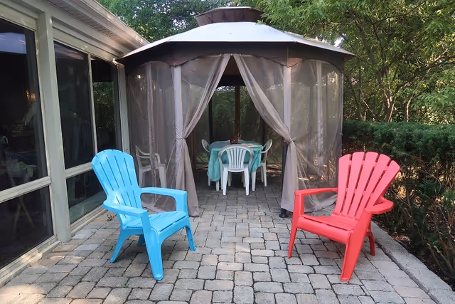 Outdoor patio area with a stone tile floor, featuring a blue plastic chair on the left and a red plastic chair on the right. In the background, there is a gazebo with sheer curtains tied back, containing a table covered with a green tablecloth and surrounded by white plastic chairs. The area is bordered by greenery and trees.