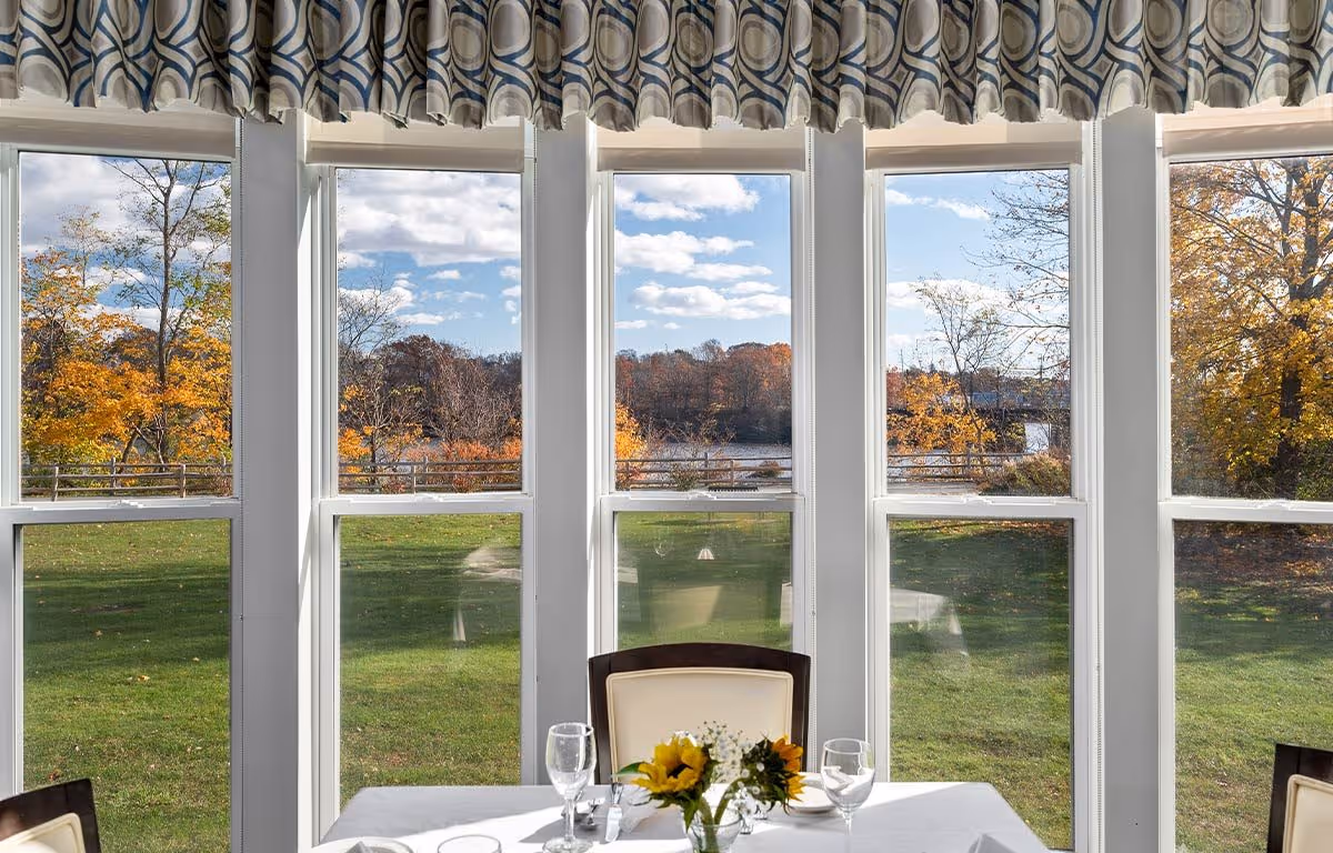 A dining table set with glassware and a small vase of sunflowers in front of large windows showing a scenic view of green grass, trees with autumn foliage, and a partly cloudy sky.