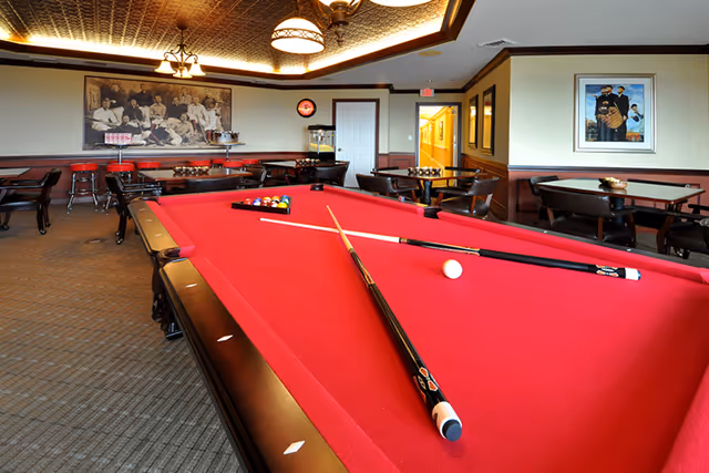 Interior view of a recreational room featuring a red pool table with two pool cues and a white cue ball on it. The room has several tables and chairs along the walls, framed artwork, a popcorn machine, and warm lighting from ceiling fixtures.