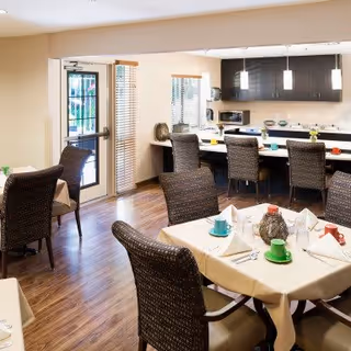 A dining area in a senior living facility with tables covered in beige tablecloths, set with colorful cups, plates, and napkins. The room has wooden flooring, wicker-style chairs, large windows with blinds, and a kitchen area with dark cabinets and pendant lights in the background.