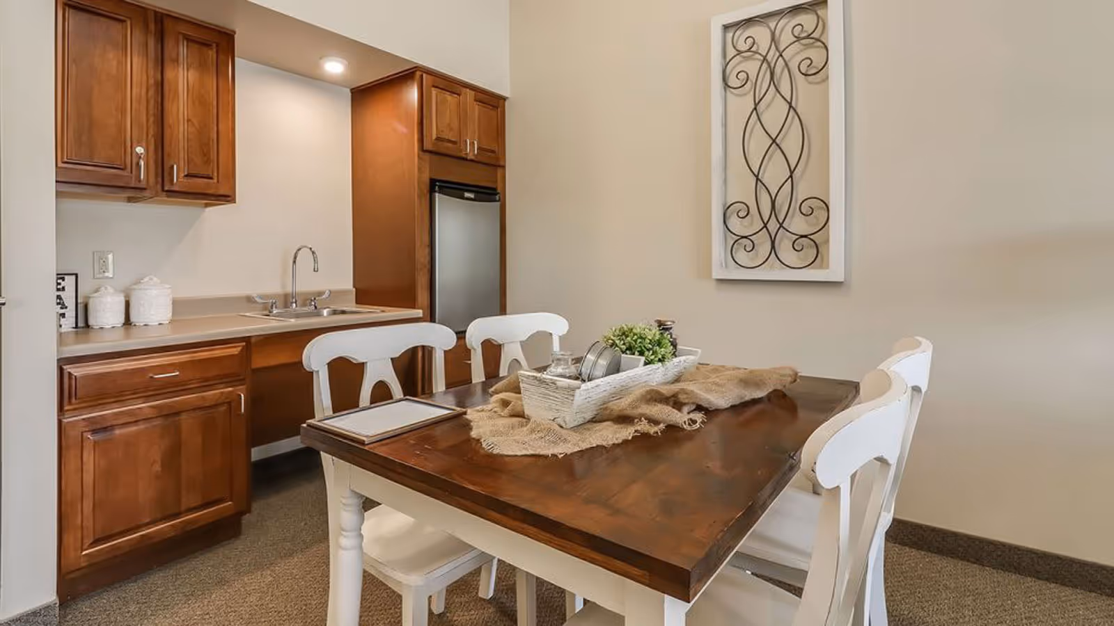 A small dining area with a wooden table and four white chairs. The table has a burlap runner and a white decorative tray with plants and jars. In the background, there is a kitchenette with wooden cabinets, a sink, and a small refrigerator. A decorative metal wall art piece is hung on the beige wall.
