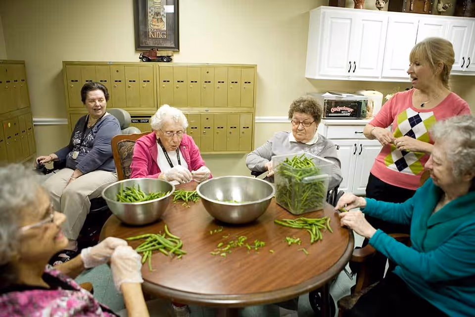 A group of elderly women sitting around a round wooden table shelling green beans into metal bowls and a plastic container in a room with mailboxes on the wall and white cabinets. One woman is standing and appears to be talking or instructing the group.