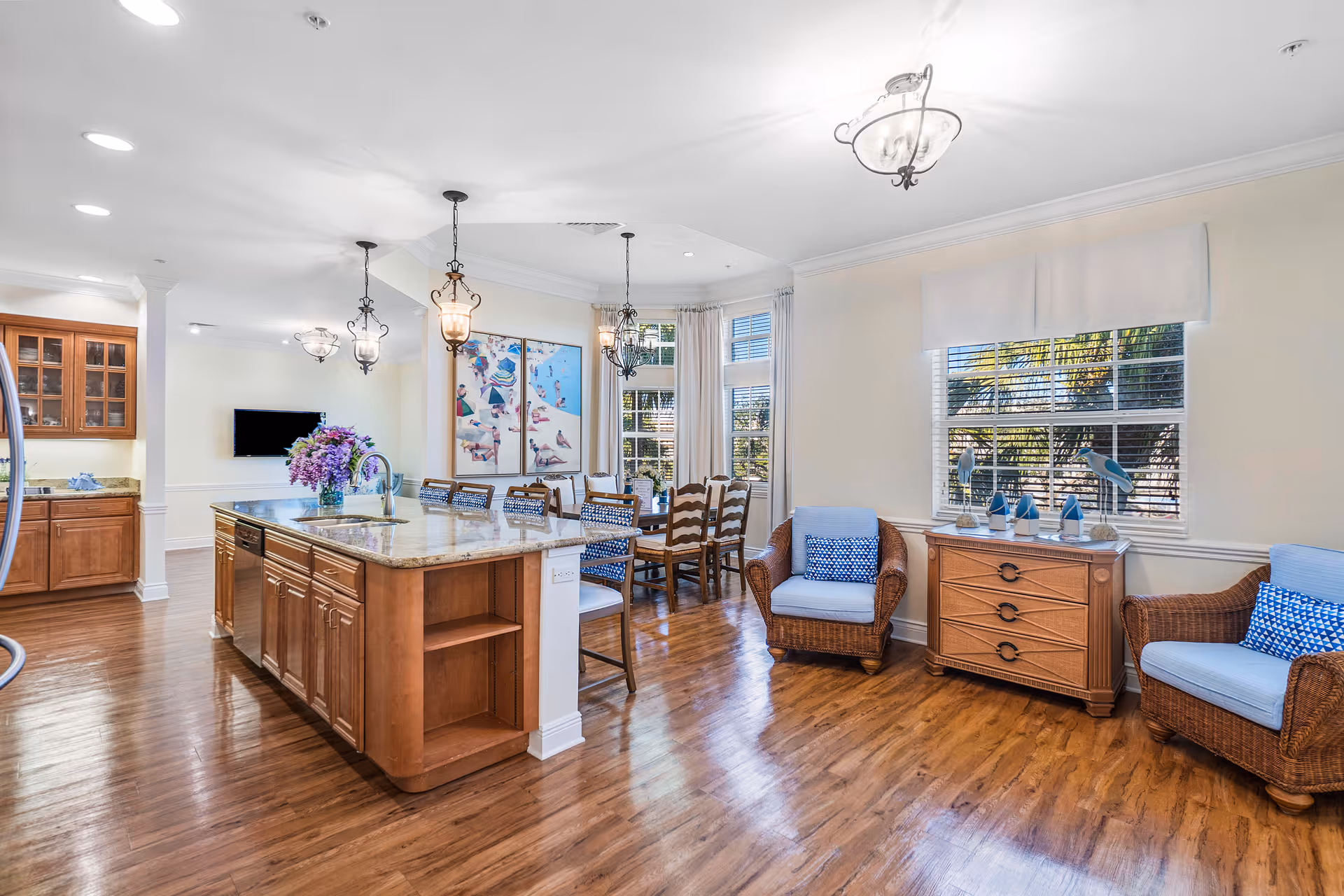 Bright and spacious interior of a senior living facility featuring a kitchen island with a granite countertop and a sink, wooden cabinets, and pendant lighting. Adjacent to the kitchen is a dining area with a wooden table and chairs, and a sitting area with two wicker armchairs with blue cushions and a wooden chest of drawers beneath a window with blinds. The room has wooden flooring and light-colored walls with large windows letting in natural light.