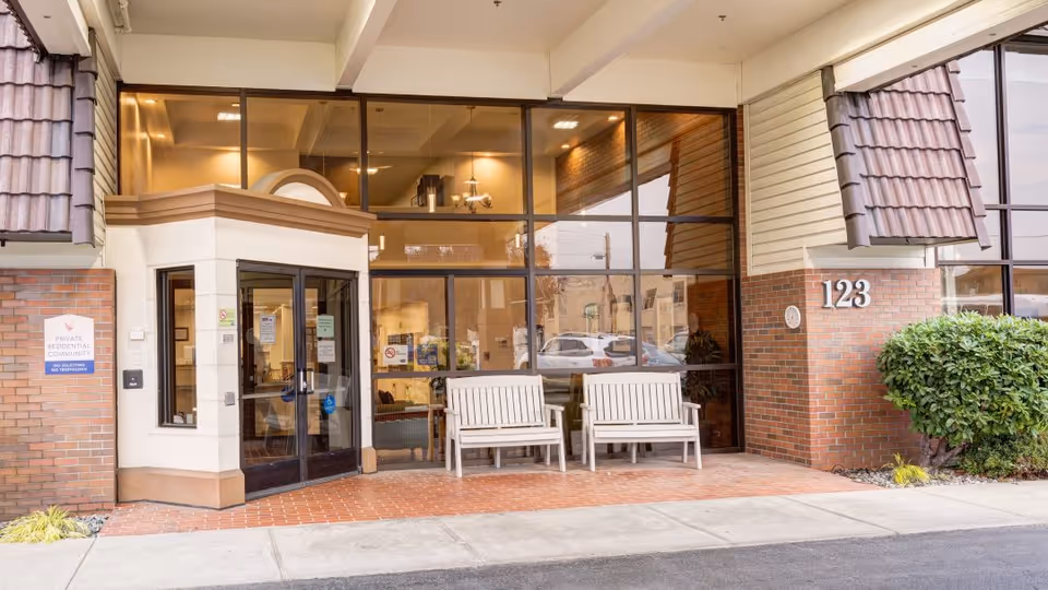 Entrance to The Rivers at Puyallup facility showing a glass door with two white benches outside on a brick patio. The building exterior features brick and beige siding with large windows above the benches. The number 123 is displayed on the brick wall to the right of the entrance.