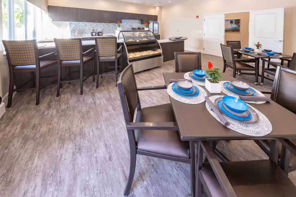 A bright dining area with several dark wood tables and chairs set with blue and gray dishware and placemats. In the background, there is a counter with high chairs and a display case containing baked goods. The room has light-colored walls and wood-look flooring.